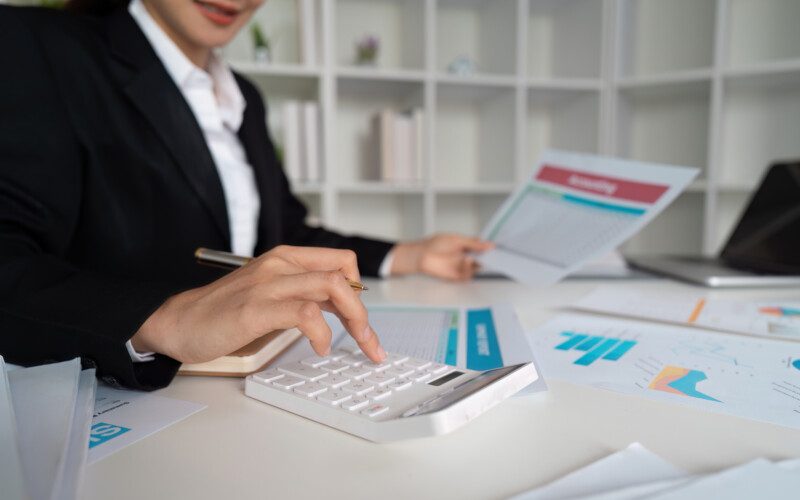 Professional businesswoman in a suit analyzing financial documents and using a calculator at a desk in a modern office.
